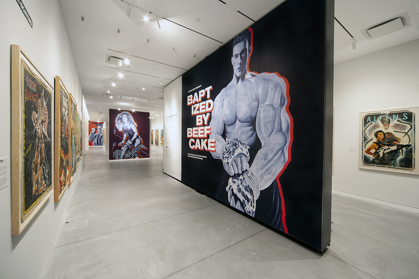 Baptized By Beefcake exhibition highlighting a larger than life, super muscled, Van Damme inlaid on a black freestanding wall.