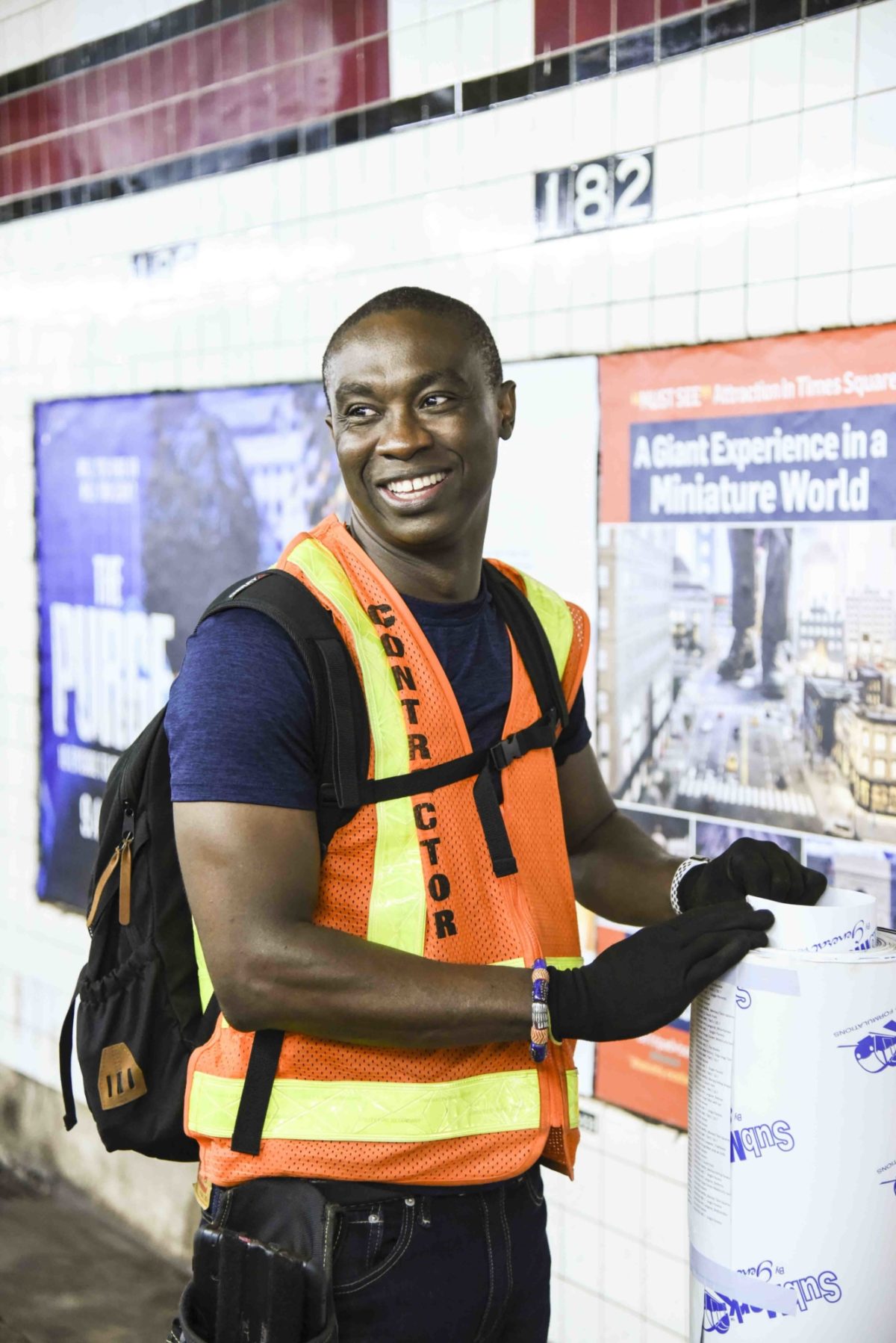 A smiling man wearing a safety best is holding a large roll of poster in a train station.
