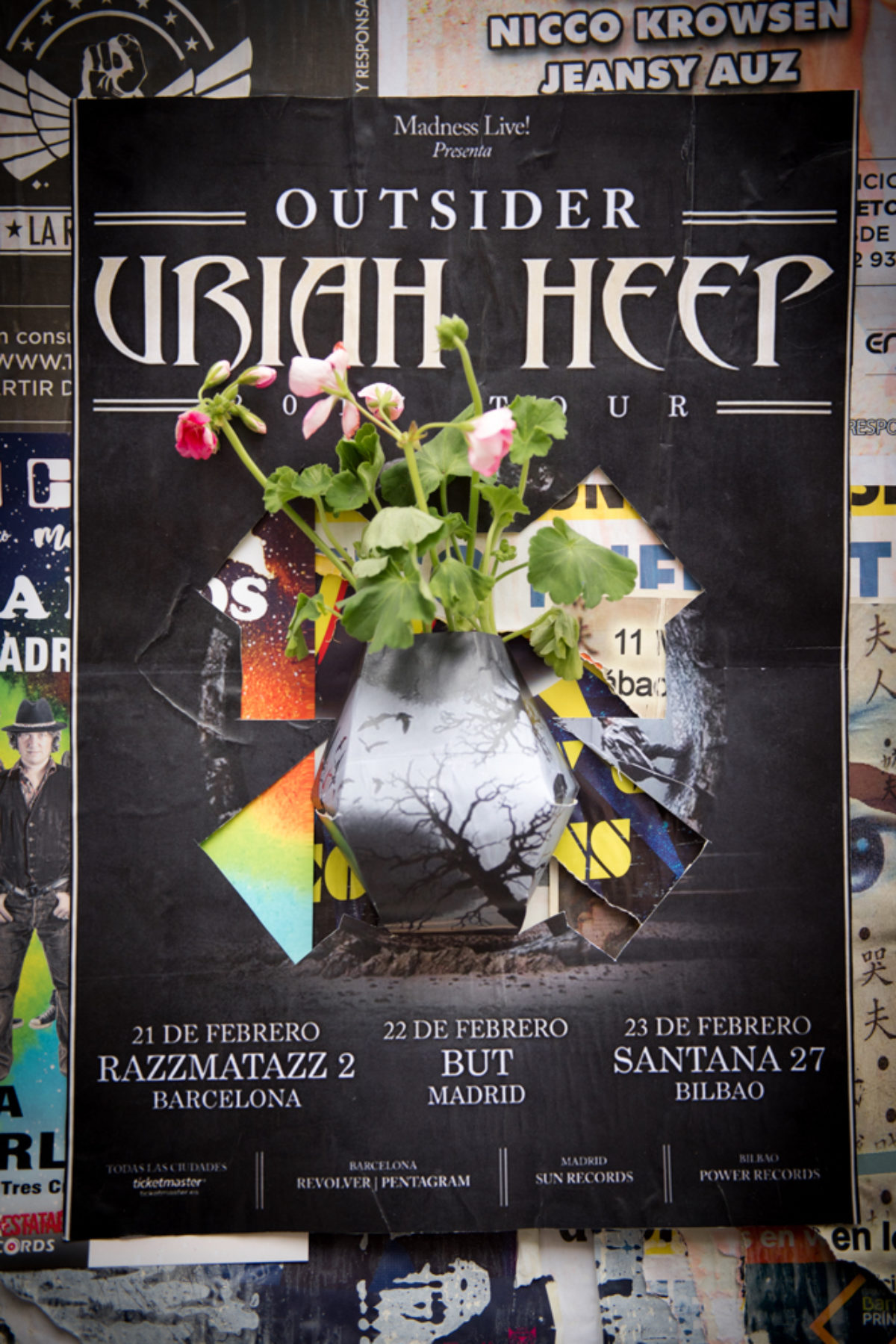 A vase with flowers are extending from a black poster advertising a concert.