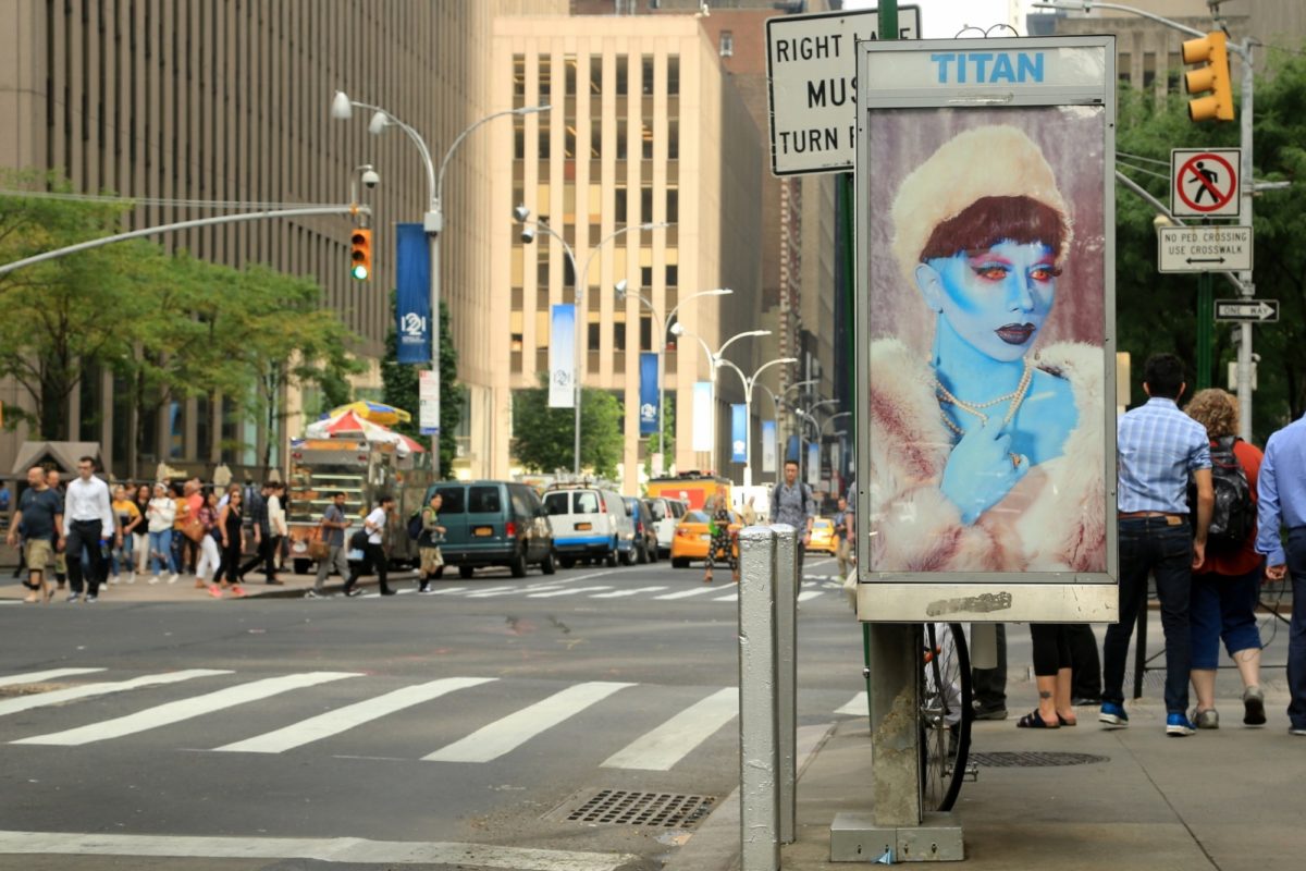 A telephone booth with a poster of a blue person at the corner of a busy New York street.