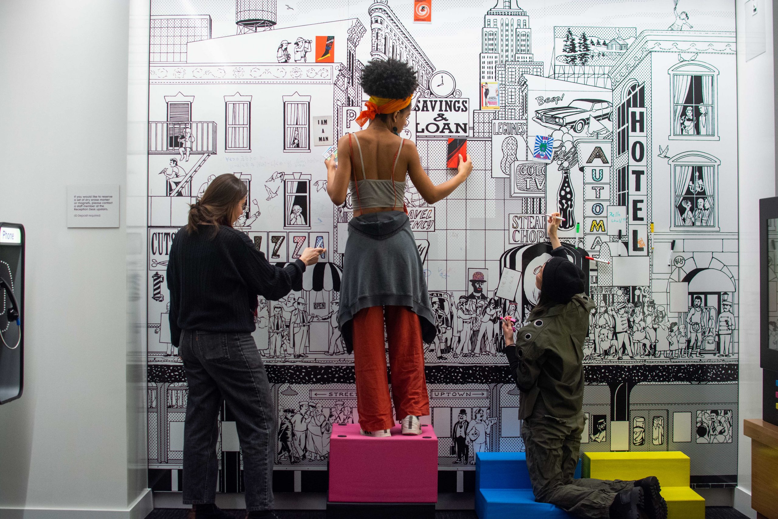 Three women adding to the 1950's inspired dry-erase board in the Children's Space at Poster House.