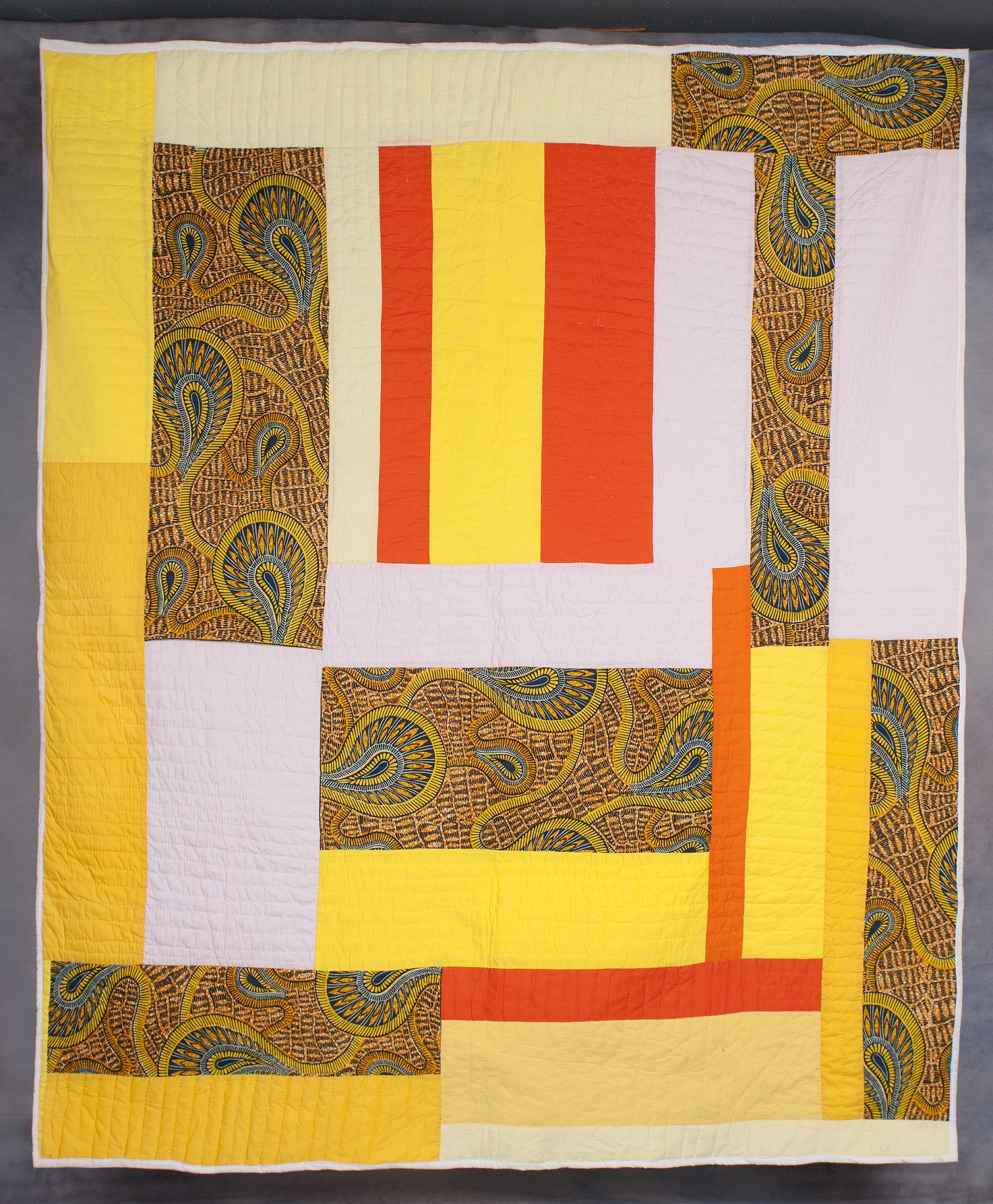 A photo of a textile made from combining various colored strips of cloth arranged in a geometric pattern with orange, yellow, white and gold paisley.