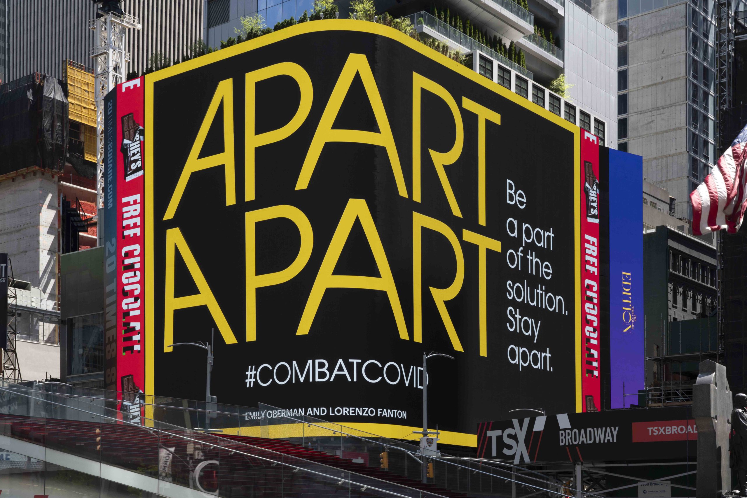 A digital billboard wrapping a corner in Times Square against a cityscape. Yellow and white text on black background. Text reads Apart Apart Be a part of the solution. Stay apart. #combatcovid Emily Oberman and Lorenzo Fanton.