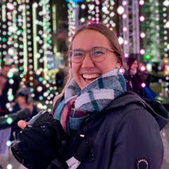 A female intern in glasses and a fluffy scarf smiling against a colored light background.