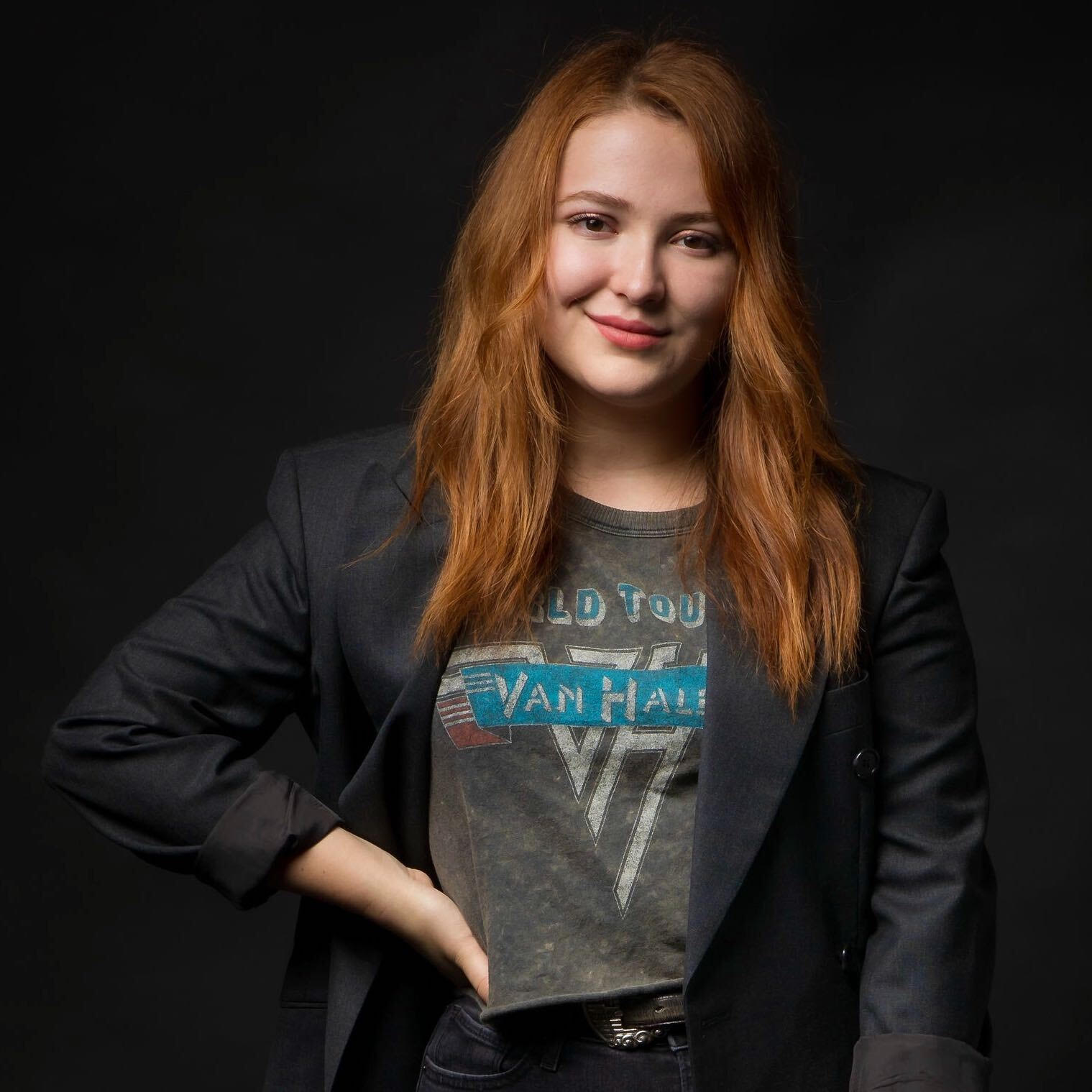 A redheaded woman in a black blazer with a hand on her hip.