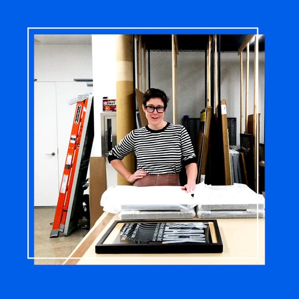 A photo of the archives room in Poster House with the Collections Manager in the center looking at the camera. The photo is framed with a blue frame.