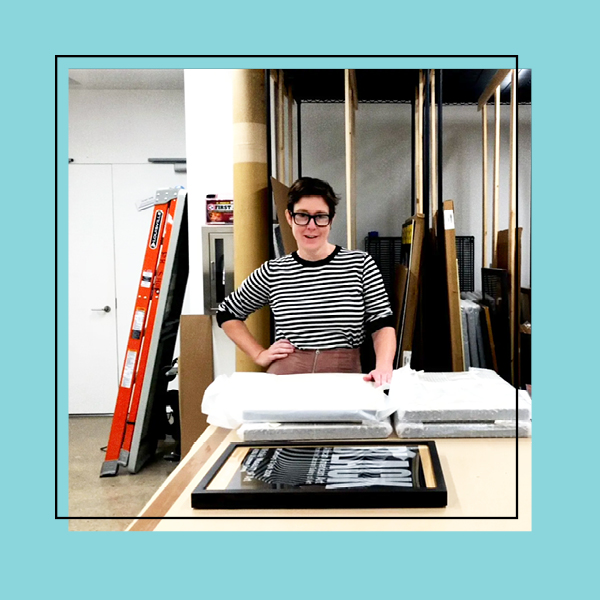 A photo of the archives room in Poster House with the Collections Manager in the center looking at the camera. The photo is framed with a teal frame.