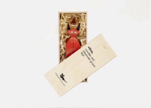 A photo featuring a red devil figurine inside a wooden casket on a gray background.