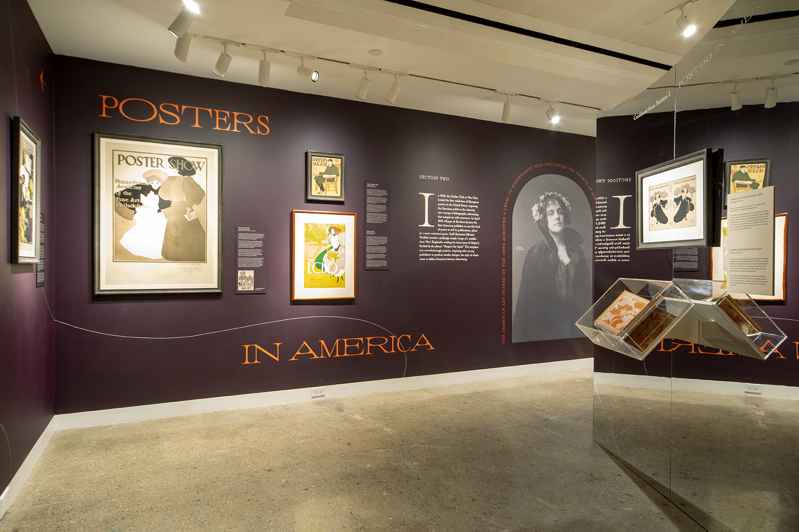 A gallery of framed posters hanging on aubergine walls next to a large black and white photo of a woman.