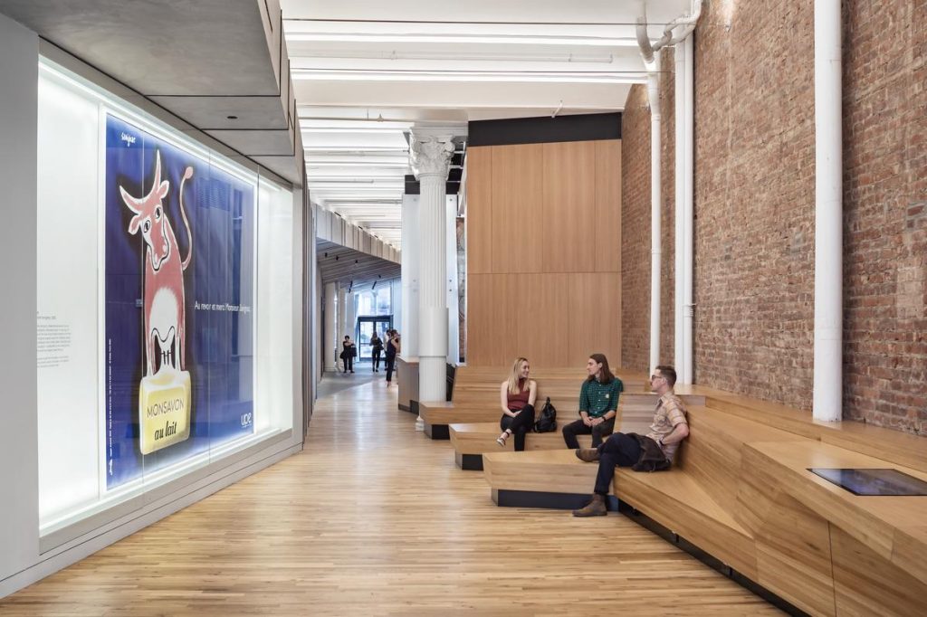 Three people sitting on angular wooden benches in the museum by a billboard of a dusty rose cow.