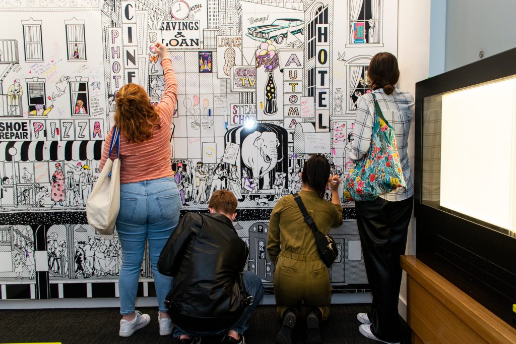 4 individuals drawing on a white board with an image of a city printed on it.
