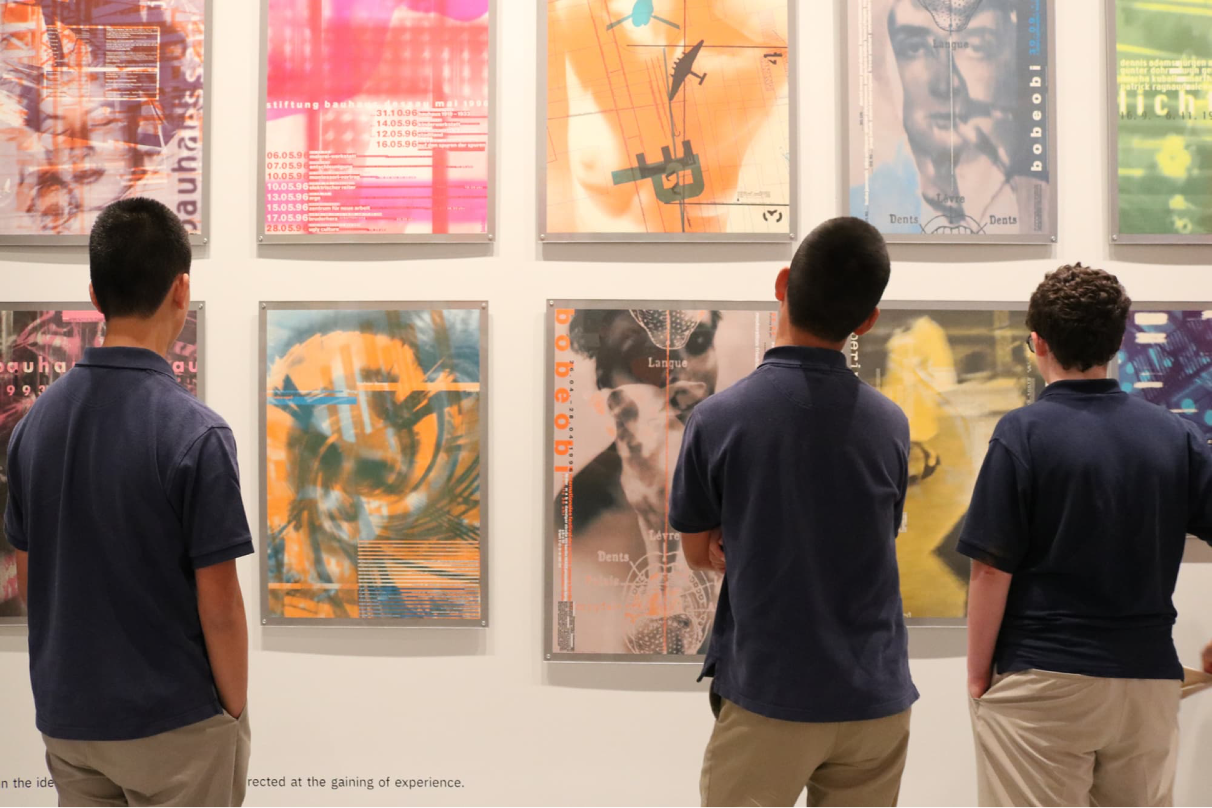 Three young people viewing ten muted toned Cyan in the 1990s exhibition framed posters on a white wall.