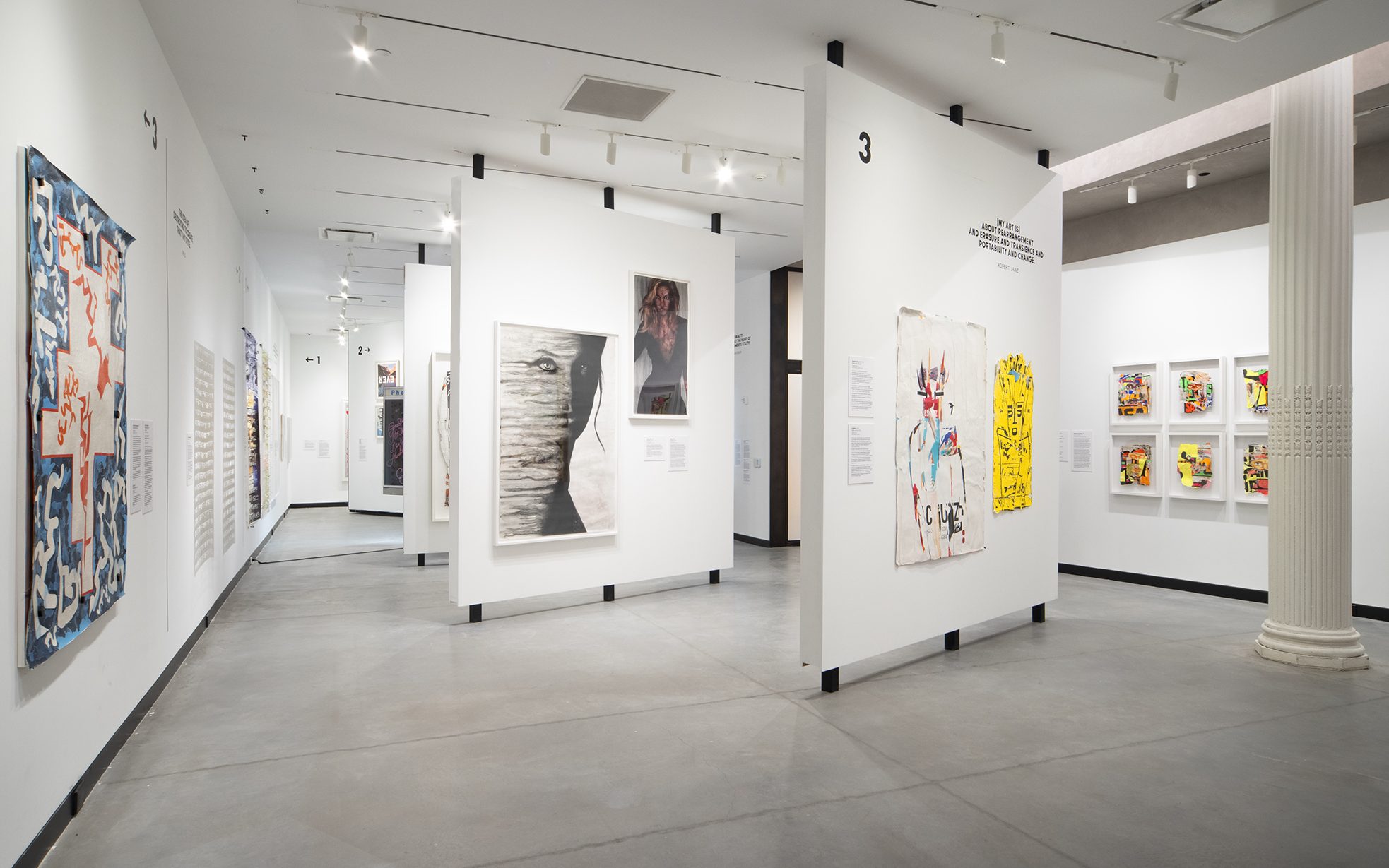 A gallery space with gray floors and white walls and white columns. Posters of mixed media styles line the outer walls and free-standing walls in the middle of the room.