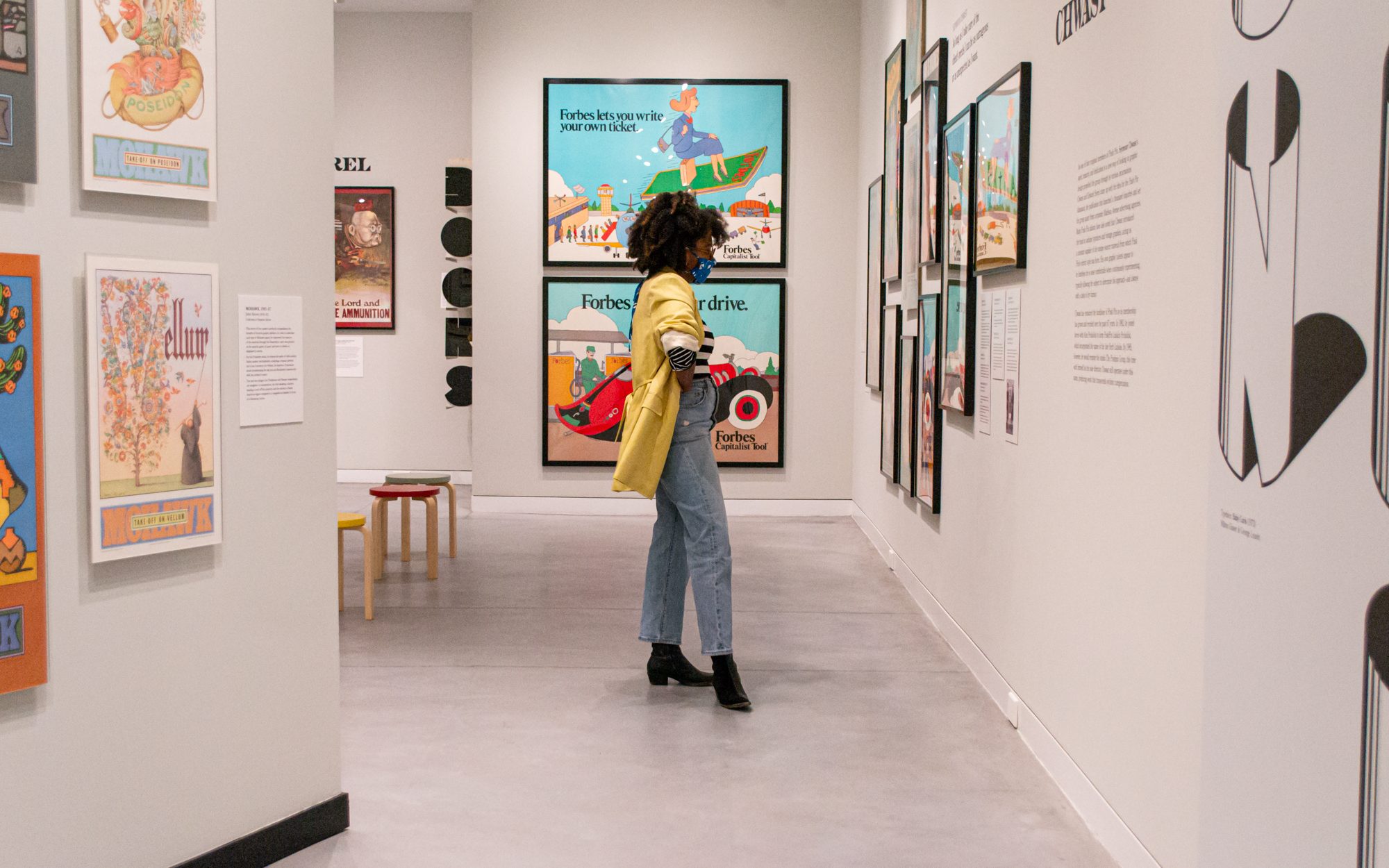 A masked figure stands in an exhibition gallery space, looking at framed posters on a wall.