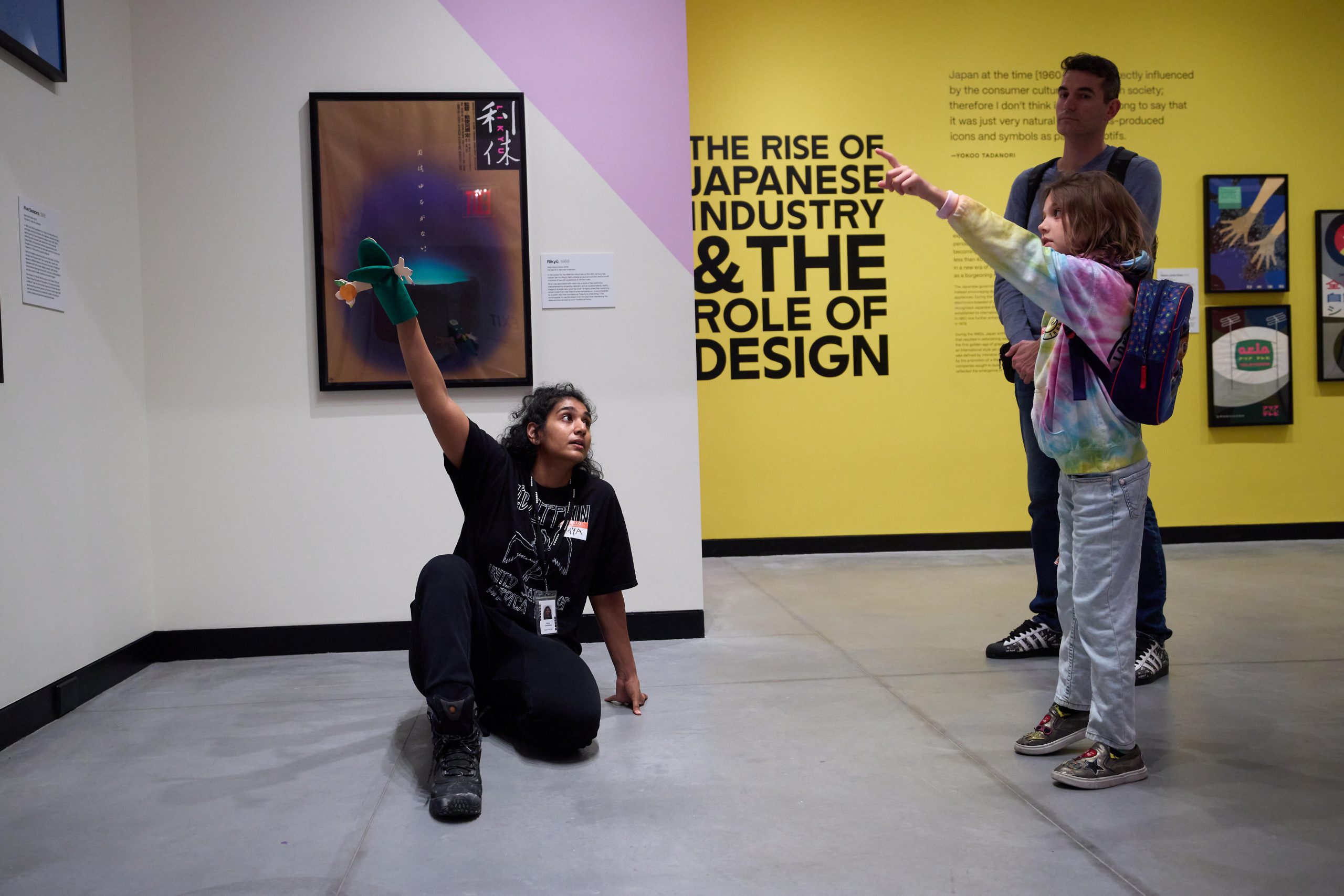 An educator with a green hand puppet kneeling in a gallery with a parent and child, who points to a poster.