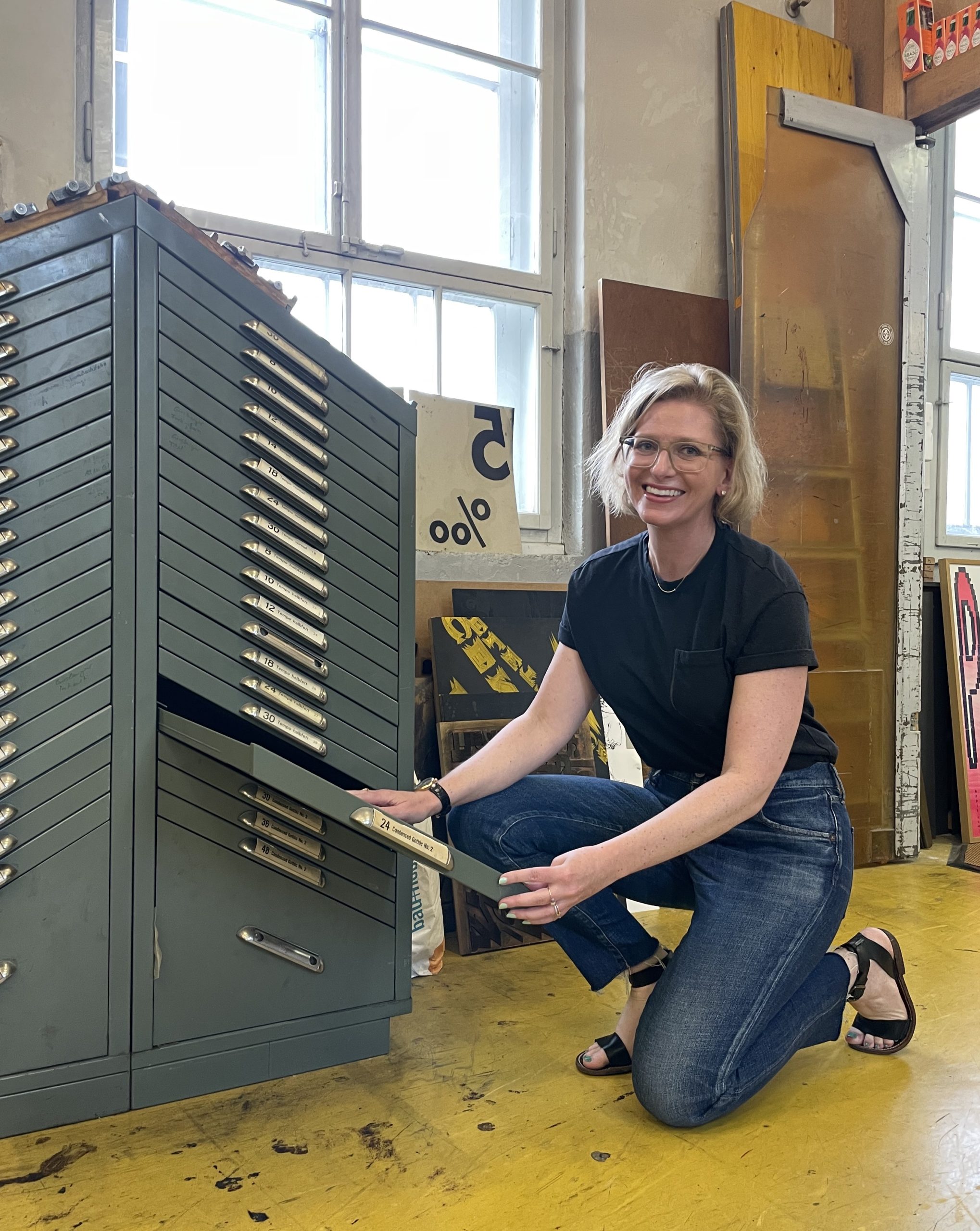 Photograph of a woman kneeling in front of a metal type case.