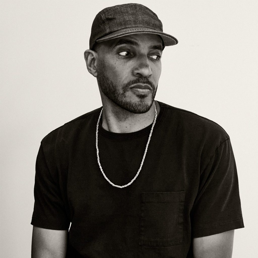A black and white photo of a Black man wearing a black t-shirt and a gold chain