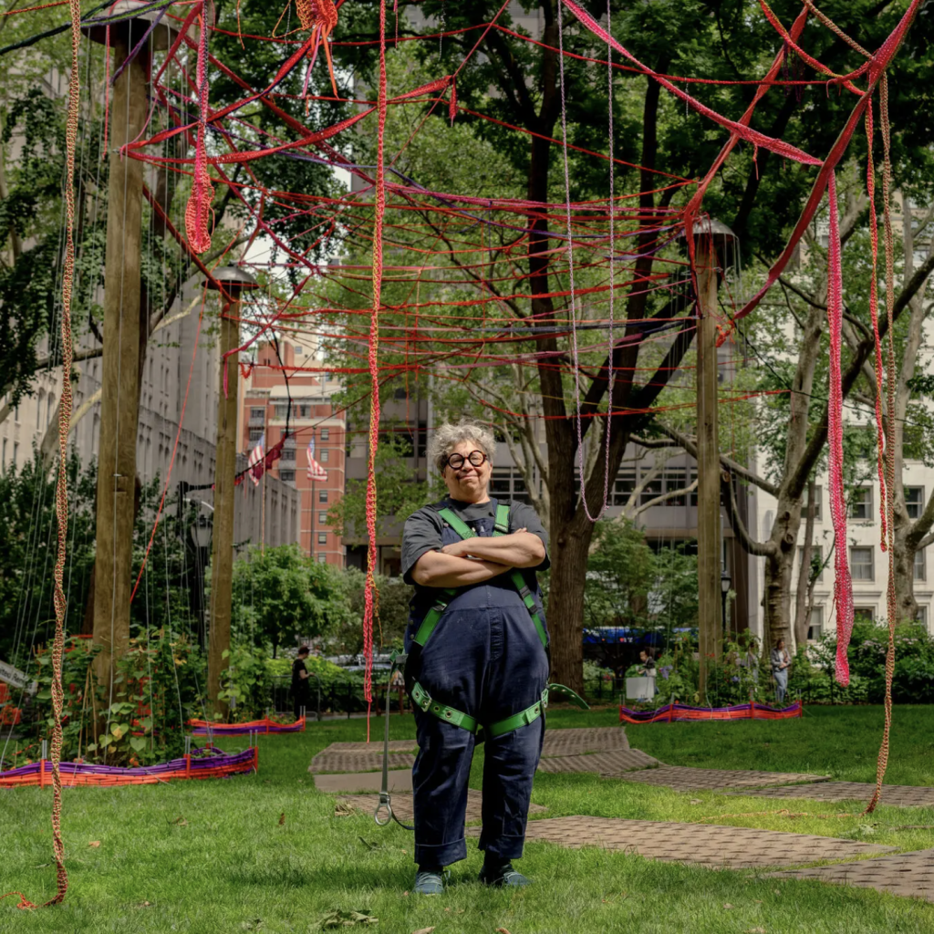 A person in a blue jumpsuit stands in front of trees covered in red strings