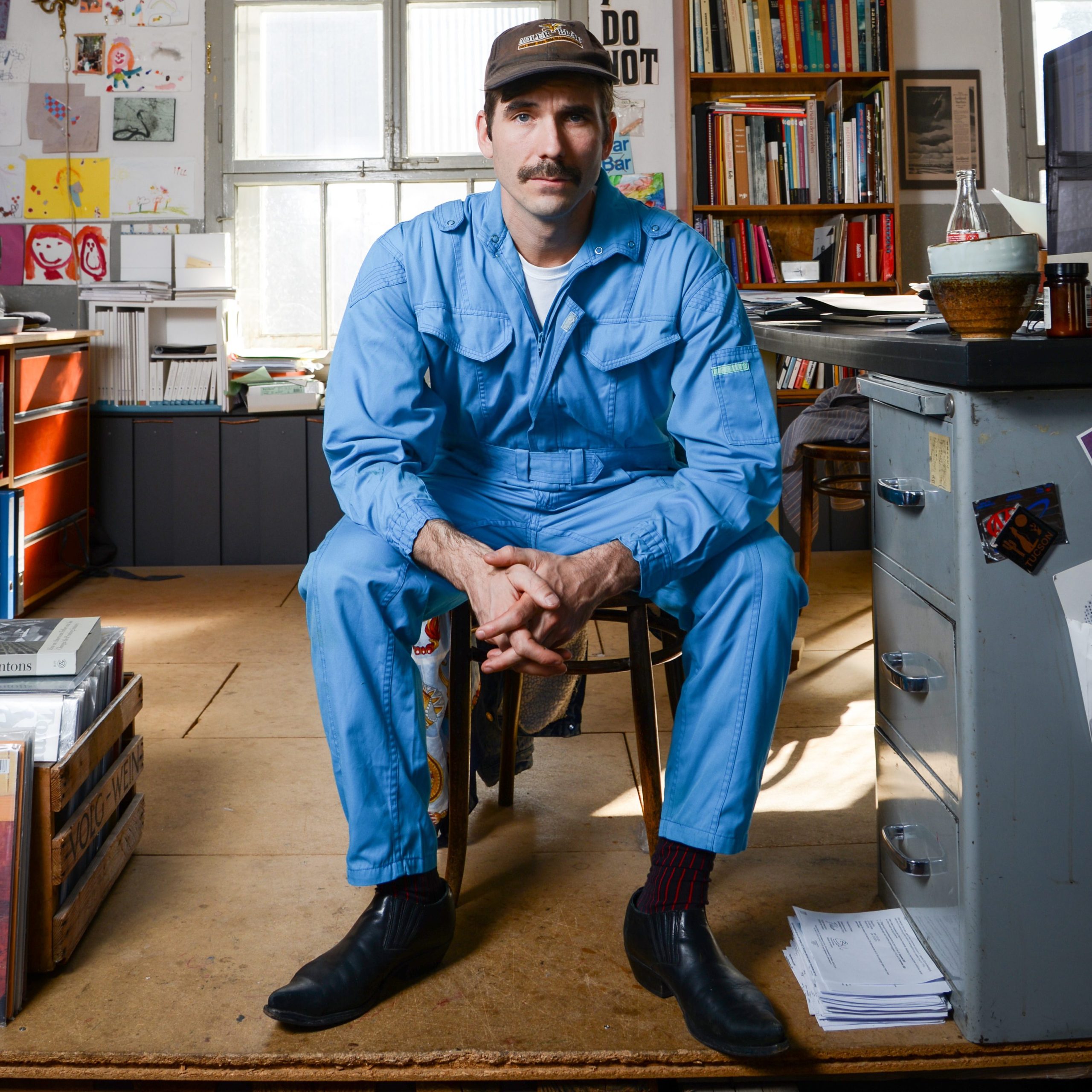 A man in a light blue jumpsuit sitting on a chair in an art studio