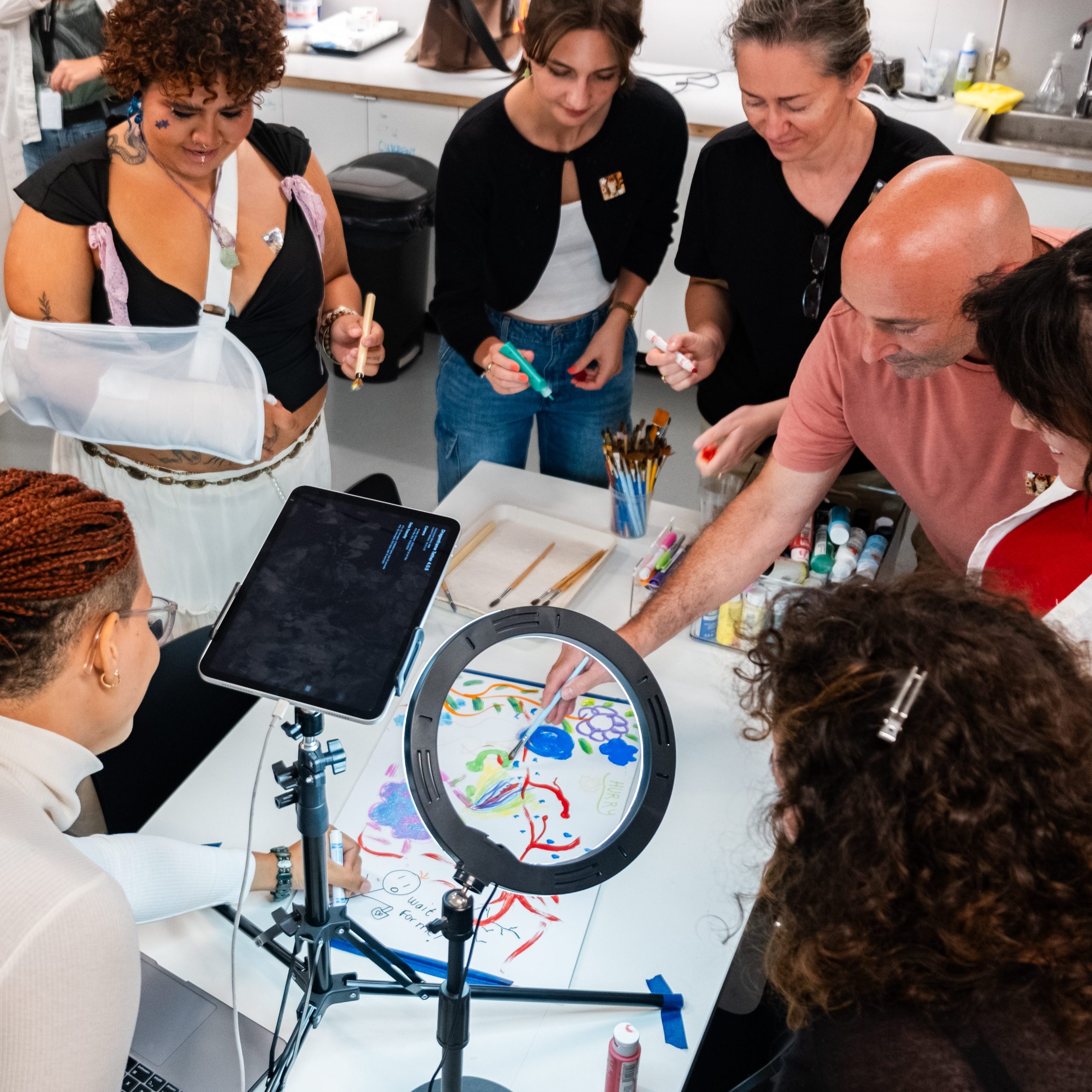 Digital image of a group of seven people standing around a table drawing on a shared piece of paper during an art workshop.