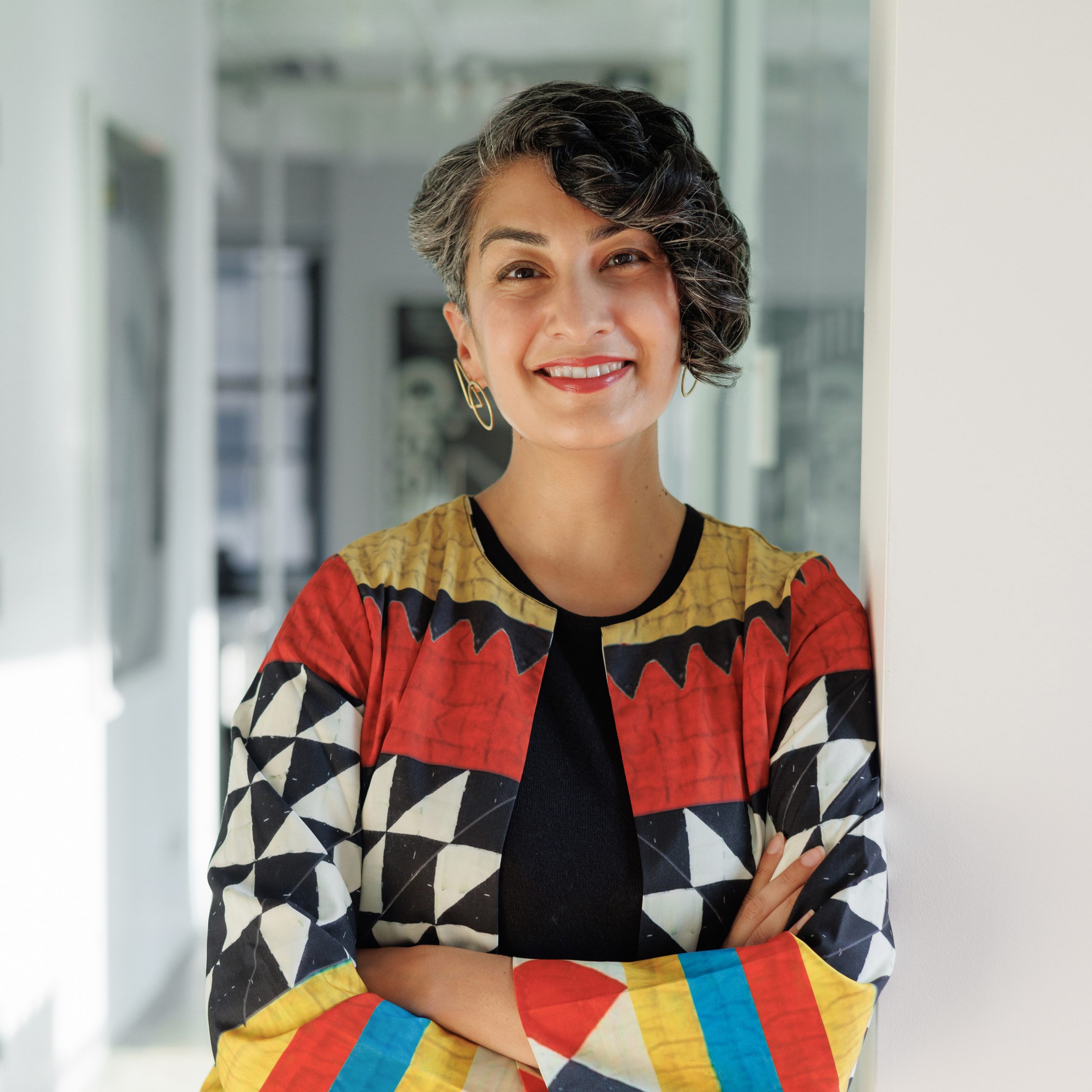 Headshot of Sara Hashim, a smiling woman with short, curled black hair wearing a colorful, geometric jacket.