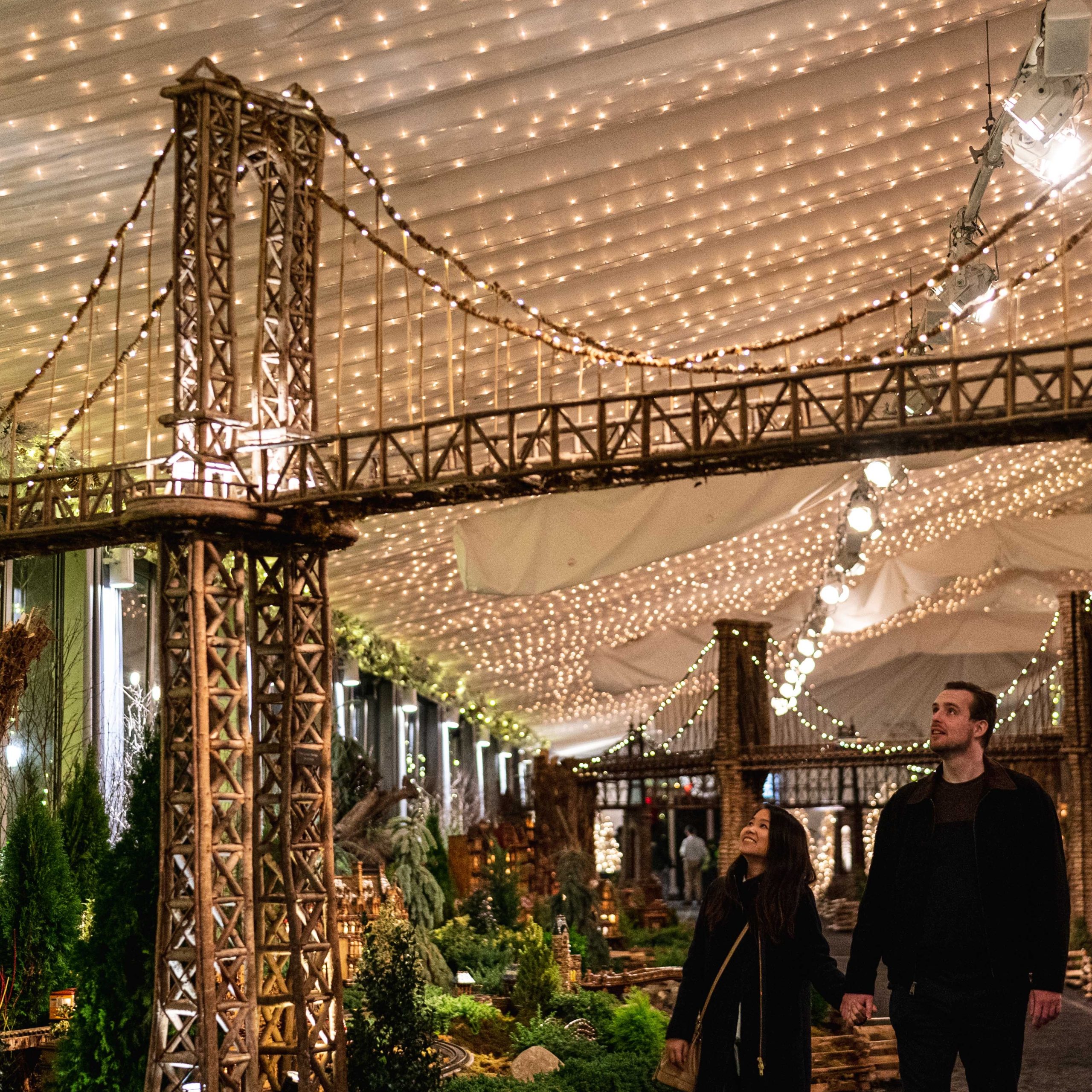 Digital image of a couple holding hands walking through a botanical garden, gazing at a statue of the George Washington Bridge, lit up by twinkling fairly lights.