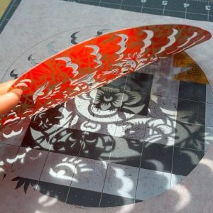 a shadow of a floral motif cast on a cutting mat by an intricately cut red piece of paper being held up to the sun