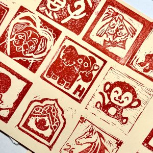 an off-white piece of paper with block printed images printed in red ink and arranged in a grid, depicting a monkey, an elephant, a dog, and other images.