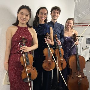 Four people standing in a line holding violins and a cello.