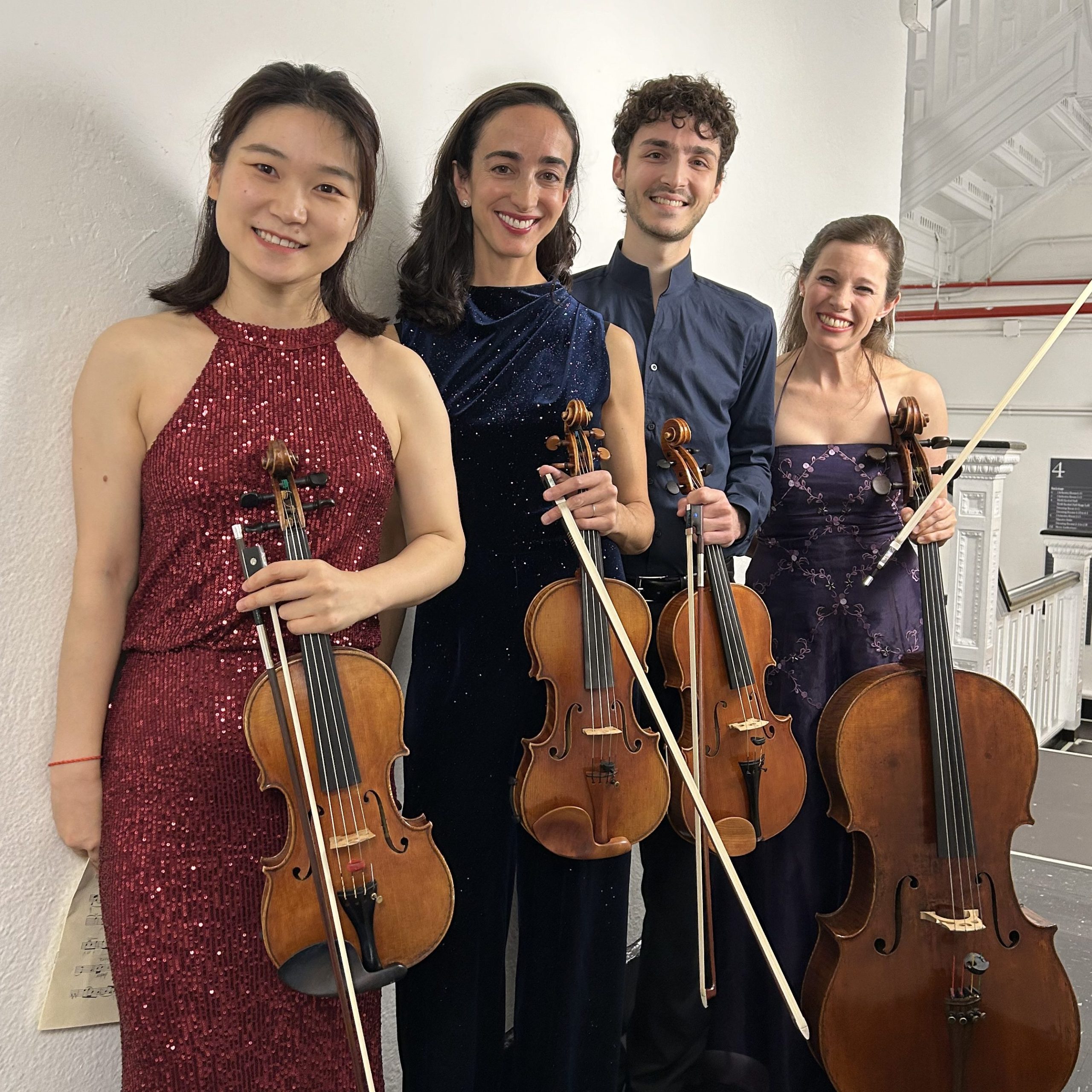 Four people standing in a line holding violins and a cello.